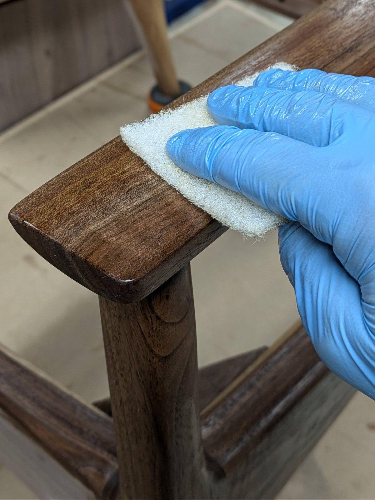 A close-up of a hand wearing a blue nitrile glove wiping a dark brown walnut chair with a white non woven pad.