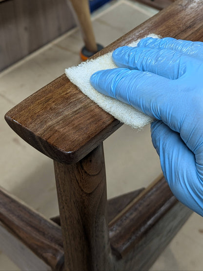 A close-up of a hand wearing a blue nitrile glove wiping a dark brown walnut chair with a white non woven pad.